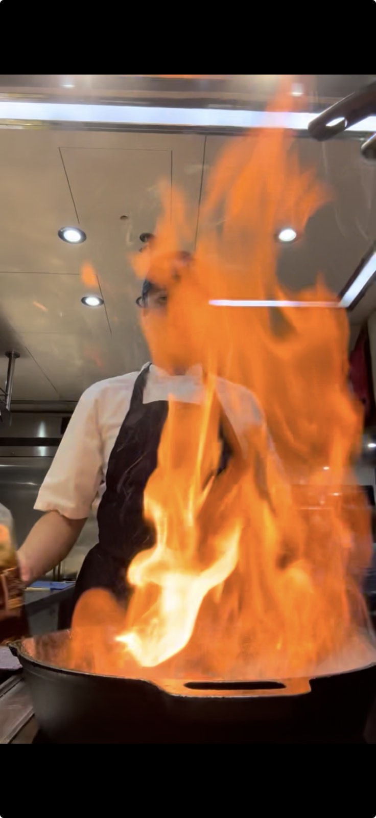 A cook behind a black pan as flame erupts from it in a professional kitchen